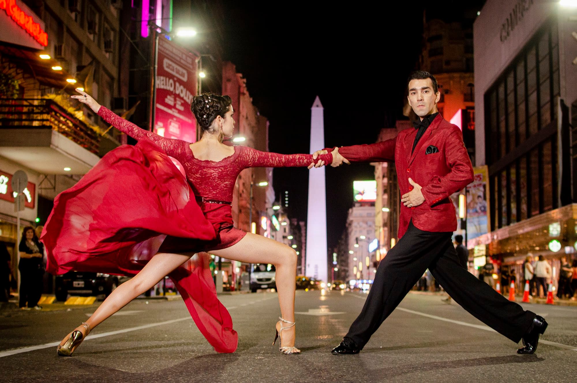 Two tango dancers in red on a Buenos Aires avenue at night, the Obelisco behind them