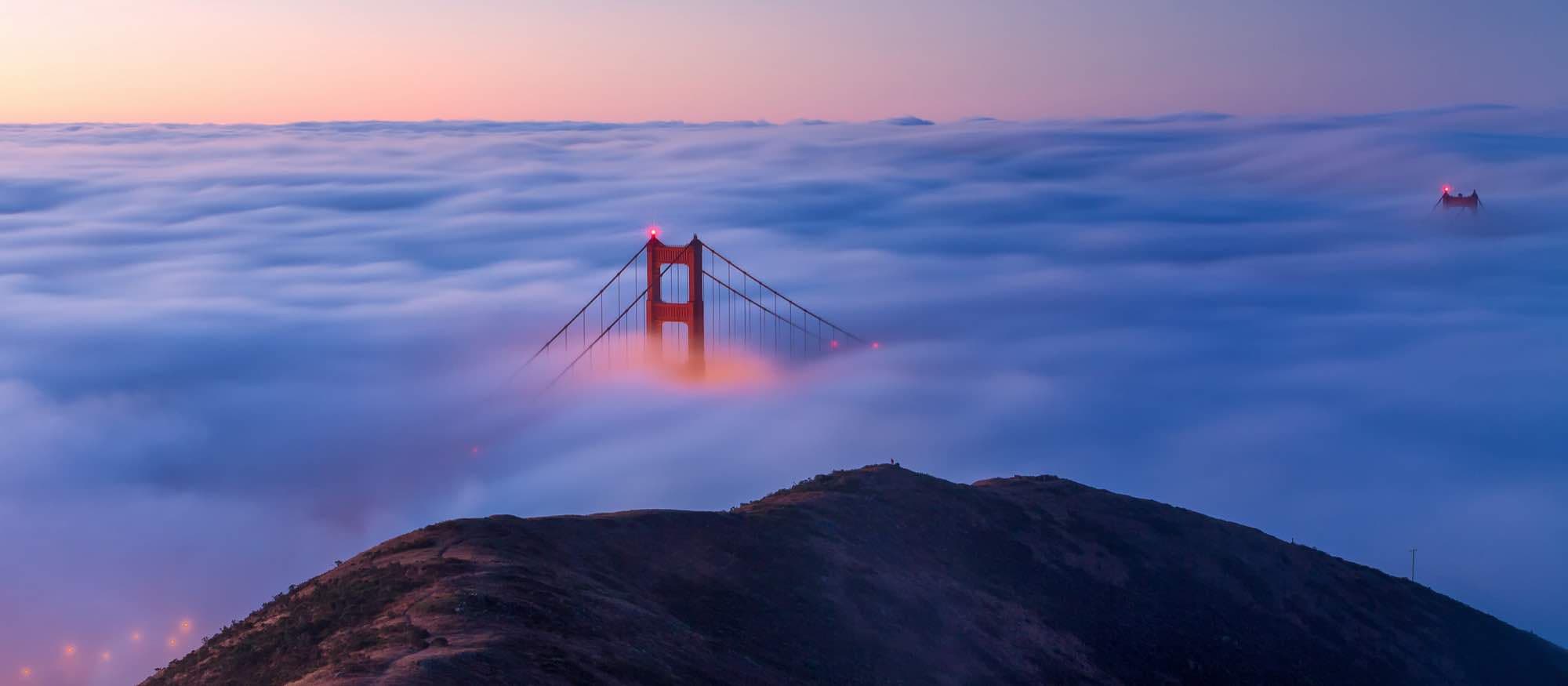 A tower of the Golden Gate Bridge rising out of pink fog at dusk